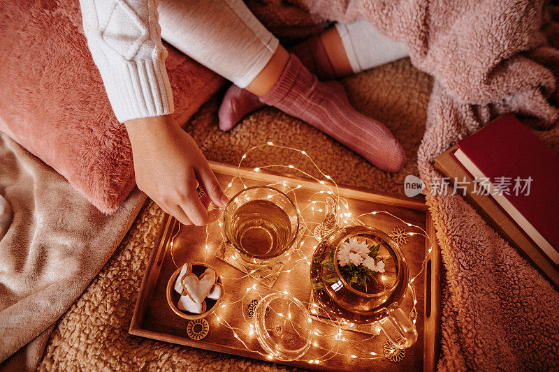 Unrecognizable young woman in cozy woolen socks on her feet and pullover with winter blankets drinking her blooming flower tea on the sofa